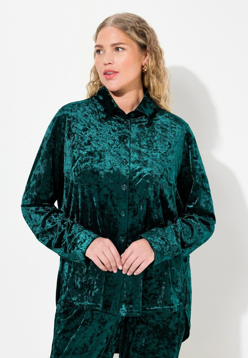 Camisa de terciopelo verde con cuello tipo polo, cierre de botones y mangas largas. La tela tiene un acabado texturizado que refleja la luz en diferentes tonalidades.