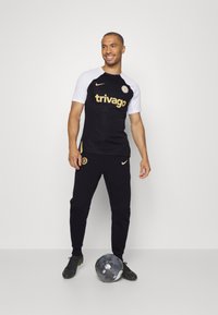 Maillot de sport noir et blanc avec un logo et un texte doré, associé à un jogging noir. Un ballon de football noir aux pieds, porté avec des crampons noirs.