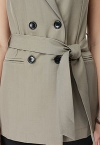 Μπεζ σακάκι διπλής σειράς με μαύρα κουμπιά και φαρδιά ζώνη δεμένη σε κόμπο στη μέση, φορεμένο πάνω από μαύρο παντελόνι.