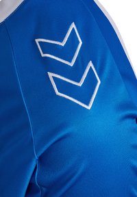 Maglia sportiva blu con texture liscia, loghi a chevron ricamati bianchi e strisce bianche a contrasto sulle spalle.