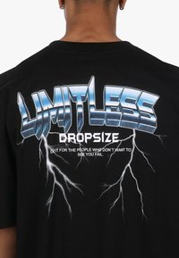 Fekete pamut póló, amelyen a "LIMITLESS DROPSIZE" kifejezés hangsúlyos grafikus nyomata látható fémes kék színben, villám díszítésekkel és fehér szöveggel.