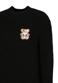 Zwarte trui van een gestructureerde stof met een tan teddybeer patch en decoratieve elementen, waaronder geldsymbolen en gekleurde accenten.
