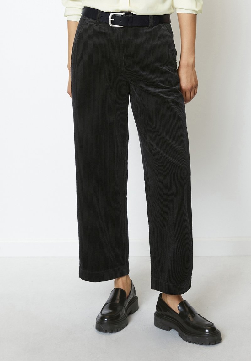 Femme portant un pantalon large en velours côtelé noir, une ceinture noire et des mocassins noirs épais, debout contre un mur blanc uni.