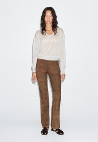 Woman with long curly hair wearing a light gray sweater, brown suede pants, and black open-toe sandals standing against a white background.