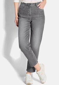 Femme portant un jean gris slim avec de petites embellissements en clous près des poches et des baskets blanches, se tenant avec les jambes croisées.