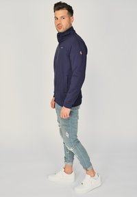 Granatowa kurtka zapinana na zamek z wysokim kołnierzem, dwiema kieszeniami bocznymi i detalem logo na rękawie; zestawiona z jasnymi dżinsami z przetarciami i białymi sneakersami.