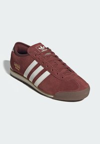 Μπορντό σουέτ αθλητικό παπούτσι με λευκό σχεδιασμό τριών ρίγων, ελαστική σόλα και χρυσή λεπτομέρεια "adidas Italia" στο πλάι. Κλασική χαμηλή σιλουέτα.