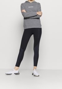 Persoon met een grijze Calvin Klein hoodie, zwart kleurblokkleggings en witte Puma sneakers met dierenprintzolen, staand met gekruiste armen.