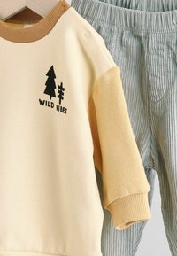 Gul sweatshirt med brun ribbet hals, featuring sort "WILD VIBES" tekst og trægrafik. Lys grøn ribbet bukser hænger ved siden af.