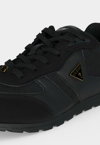 Μαύρο sneaker χαμηλού τύπου με κορδόνια, υφασμένο πέλμα και χρυσό λογότυπο Guess στο πλάι και στην άκρη των κορδονιών, εμφανίζεται σε κοντινή γωνία.