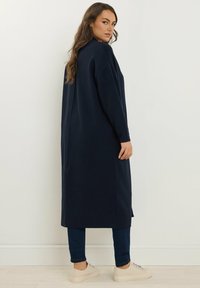 Lungo cardigan blu navy realizzato in tessuto morbido, con vestibilità comoda, tasche laterali e polsini a costine. Indossato sopra jeans scuri e scarpe chiare.
