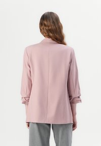 Blazer rose clair avec une coupe ajustée, des revers crantés et des manches retroussées. Le tissu semble lisse avec une silhouette structurée.