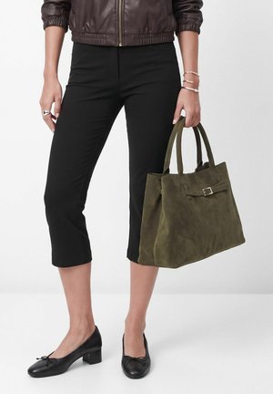 Personne portant un pantalon noir court, des chaussures noires à talons bas et une veste marron, tenant un sac à main en daim vert olive avec détail de boucle.