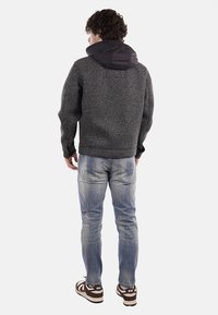 Giacca grigia con texture e cappuccio nero, abbinata a jeans blu sbiaditi e sneaker marroni e bianche. Cuciture e dettagli visibili sulla giacca.