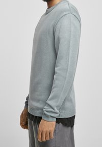 Světle šedý texturovaný svetr s kulatým výstřihem a dlouhými rukávy, s vroubkovanými manžetami a spodním lemem. Nesený přes černou košili, zobrazený z boku.