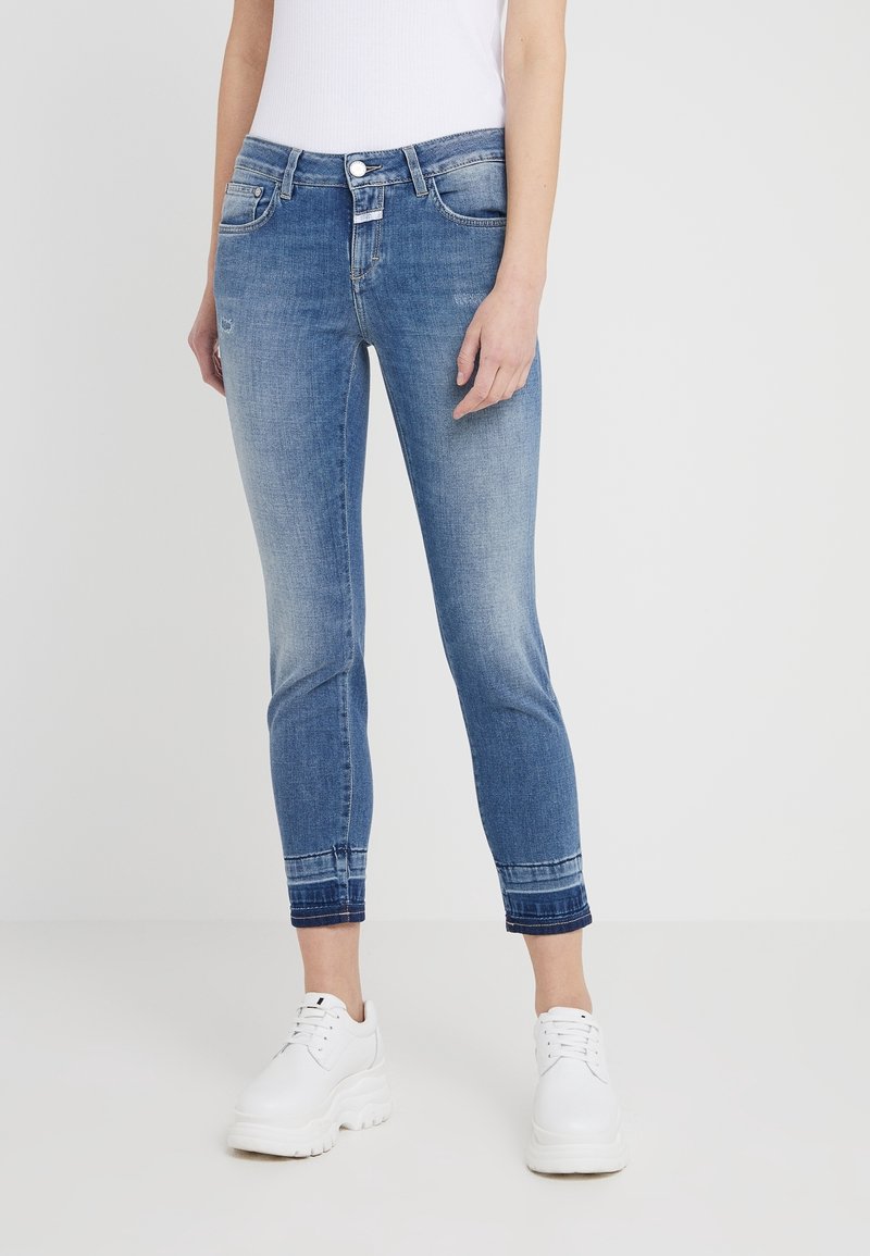 Vybledlé modré denimové džíny s slim fit, s hladkým lemem a pěti kapsami, spárované s bílými teniskami. Hladká textura a střední pas.