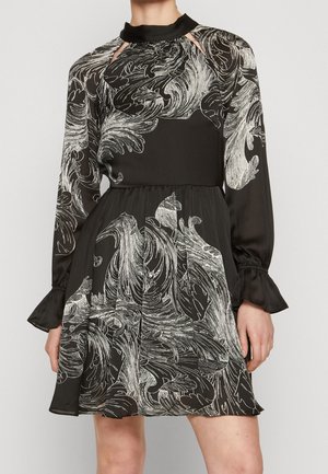 Mujer con un vestido negro con patrones florales blancos en espiral, mangas largas, puños con volantes y aberturas cerca del escote.