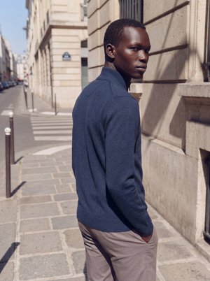 Jeune homme à la peau foncée portant un pull bleu et un pantalon beige, debout sur un trottoir en ville, les mains dans les poches, regardant par-dessus son épaule.