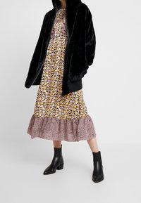 Czarna kurtka z sztucznego futra zapinana na zamek, noszona na kwiecistej sukience z żółtymi i białymi kwiatami, falbaną na dole oraz ciemnymi akcentami w zwierzęcy wzór. Czarne botki za kostkę.