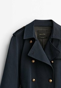 Manteau croisé bleu marine foncé avec boutons dorés, larges revers et épaulettes, suspendu sur un fond blanc.