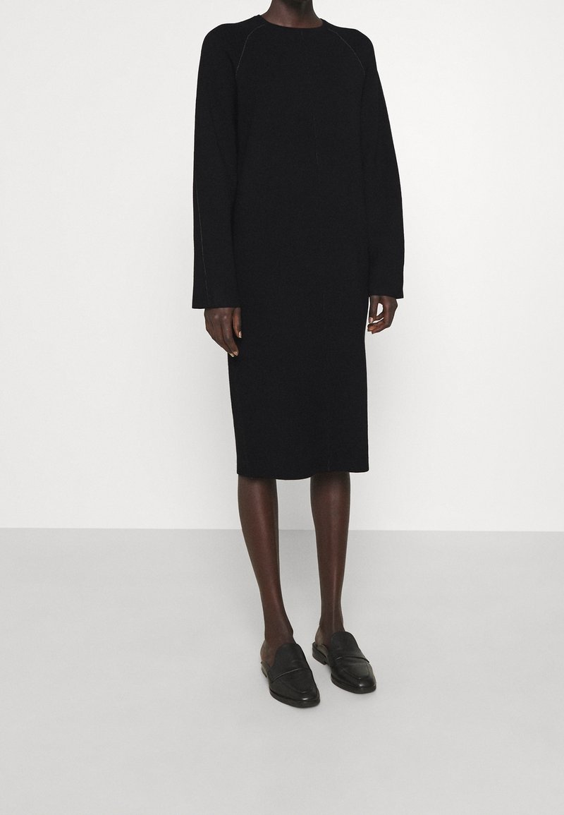 Robe noire à manches longues avec une silhouette droite et un ourlet mi-genoux, dotée d'un col rond et de détails de couture subtils. Assortie à des mocassins noirs.
