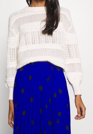 Femme portant un pull blanc en maille à manches longues et une jupe plissée bleue avec de gros pois noirs.