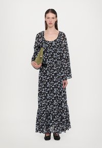 Abito lungo nero floreale con fiori blu e bianchi, tessuto trasparente, orlo a ruches, abbinato a una borsa verde e scarpe nere.