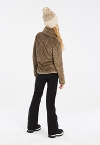 Giacca in pile marrone e morbida con colletto alto, indossata con pantaloni neri e stivali. Il cappello è color crema con un pompon sulla sommità.