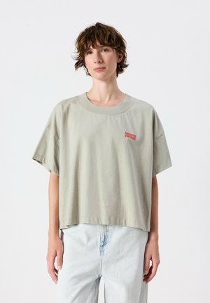 Persona con capelli corti e ricci che indossa una maglietta oversize grigio chiaro con logo rosso "American Vintage" e jeans azzurri, in piedi contro uno sfondo bianco.
