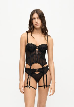 Femme debout portant un ensemble de lingerie noire en dentelle comprenant un corset, une jarretière et une culotte à bretelles sur fond blanc.