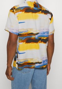 Man med kortärmad skjorta med abstrakta mönster i gult, blått, svart och orange samt ljusblå denimjeans, rygg mot kameran.