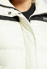 Biała kurtka puchowa z czarnymi akcentami. Posiada zamek błyskawiczny z przodu oraz zatrzask, teksturowaną tkaninę i dwie duże kieszenie z przodu.