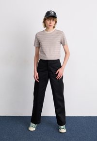 Stribet beige t-shirt med korte ærmer, sorte brede bukser og grønne sneakers. Sort kasket med logo, ensfarvet baggrund.