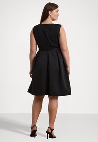 Svart ärmlös klänning med en figursydd överdel och utsvängd kjol. Har en dragkedja i ryggen och en bred halsringning. Kombinerad med svarta klacksandaler.