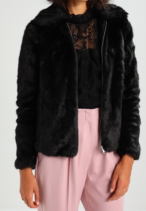 Giacca in ecopelliccia nera con chiusura a zip, dotata di cappuccio e texture morbida. Indossata con una maglia nera in pizzo e pantaloni rosa chiaro.