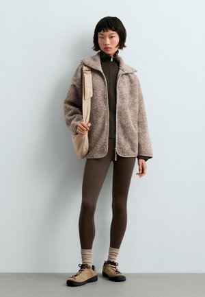 Jeune personne portant des leggings marron, une veste polaire beige, des chaussettes beiges et des chaussures de randonnée couleur cuir clair, portant un sac à bandoulière beige sur un fond uni.