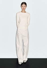 Vrouw in een crème kleurige langemouwen top en witte wijde broek, staande met handen achter de rug tegen een effen lichte achtergrond.