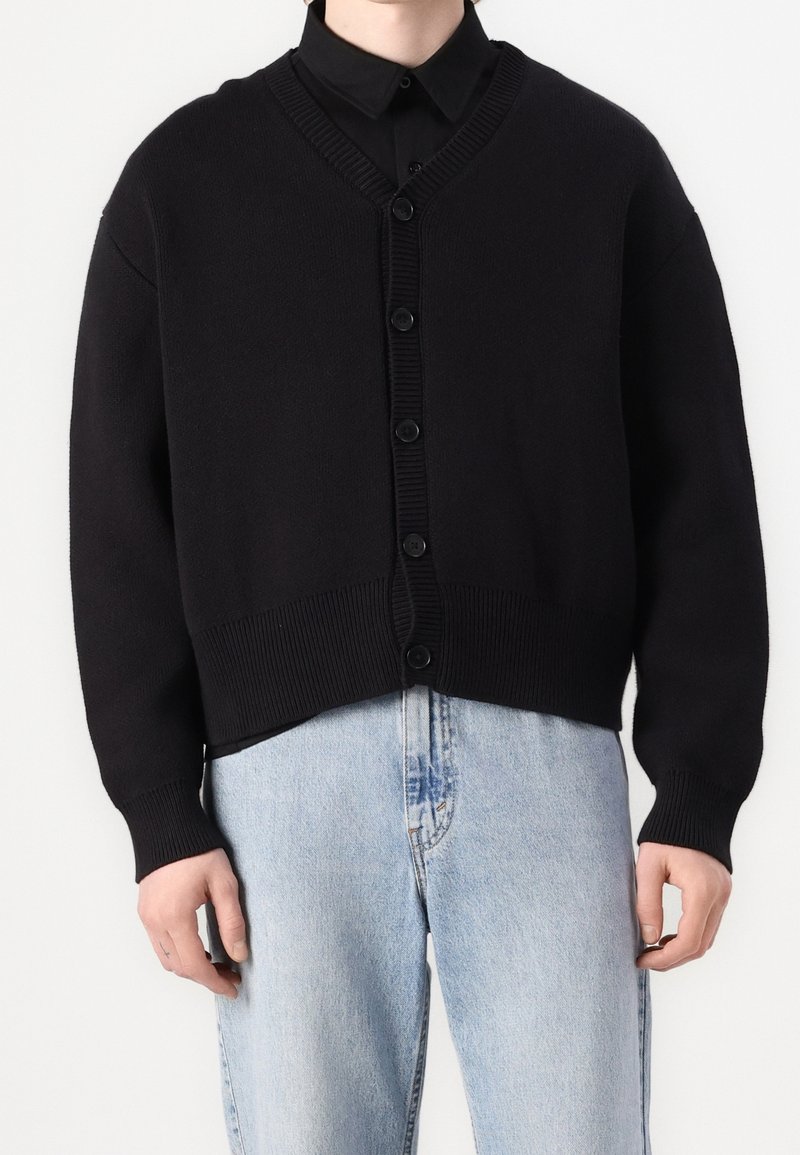Personne portant un cardigan noir boutonné sur une chemise noire à col et un jean bleu clair, debout devant un fond blanc.