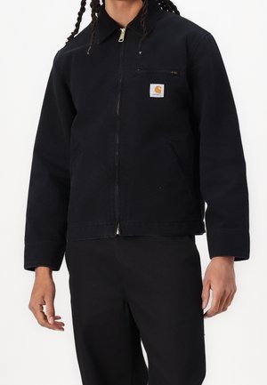 Personne portant une veste noire zippée avec une poche poitrine et un patch logo Carhartt beige, associée à un pantalon noir sur fond blanc.