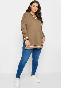 Top de manga larga marrón con estampado y escote en V, con acentos a rayas en negro y blanco, combinado con vaqueros ajustados azules y zapatillas blancas.