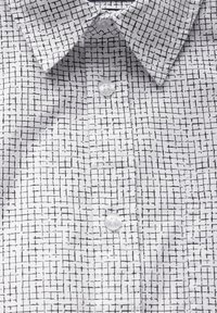 Bílá látková košile s černou mřížkovou potiskem, klasickým límečkem a předním zapínáním na průhledné knoflíky.