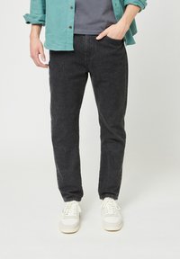Uomo che indossa jeans denim neri, sneaker bianche, maglietta grigia e camicia azzurro turchese con le maniche arrotolate, in piedi contro uno sfondo bianco.