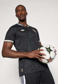 Must khašasärge, millel on lilleline tekstuur, rohe- ja valge triibuline krae, Adidas'i logo ja hem-tag. Mudel hoiab jalgpalli.