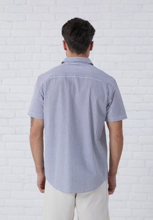 Homme de dos portant une chemise à manches courtes à rayures verticales bleues et blanches et un pantalon de couleur claire devant un mur en briques blanches.