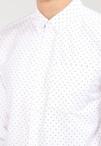 Bílá košile na knoflíky s malým, opakujícím se černým vzorem. Má náprsní kapsu a klasický límec.