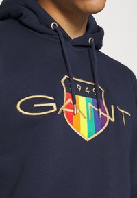Sweat-shirt à capuche bleu marine en coton, orné d'un logo brodé doré avec un écusson aux couleurs de l'arc-en-ciel et des accents de cordon.