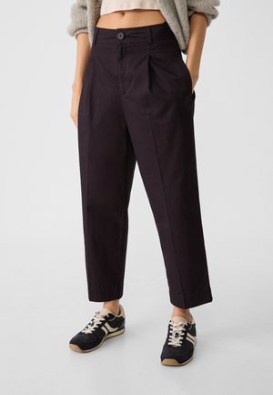 Personne portant un pantalon noir taille haute fuselé, un pull court et des baskets noires à lacets blancs, debout les mains dans les poches.
