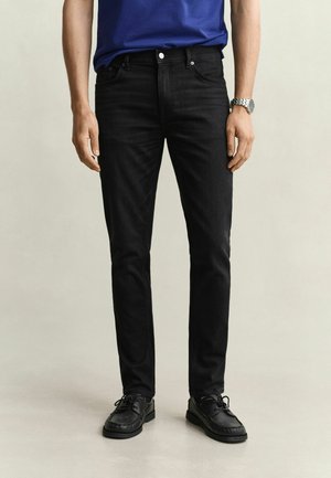 Man wearing slim-fit black jeans, black leather shoes, a blue shirt, and a silver wristwatch standing against a light background.