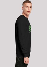 Svart langermet sweatshirt med grønn tekstdesign, rund hals, ribbestrikkede mansjetter og liv. Modellen poserer i profil mot en nøytral bakgrunn.