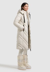 Lungo cappotto imbottito beige con motivo a zigzag, cappuccio, dettagli neri della cerniera e tasche laterali, abbinato a stivaletti beige fino al ginocchio.